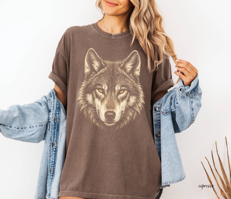 Puede incluir: Una camiseta marr&oacute;n con una ilustraci&oacute;n detallada de la cara de un lobo con ojos amarillos. La camiseta tiene un aspecto vintage y desgastado. La palabra "espresso" est&aacute; impresa en la esquina inferior derecha. Una chaqueta vaquera azul claro est&aacute; sobre la camiseta.