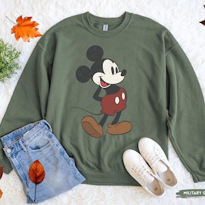 Puede incluir: Sudadera verde militar con un dise&ntilde;o de Mickey Mouse de estilo vintage. La parte superior de manga larga se combina con vaqueros azules y zapatillas blancas, creando un look casual. Hojas de oto&ntilde;o y una peque&ntilde;a planta a&ntilde;aden acentos de temporada.