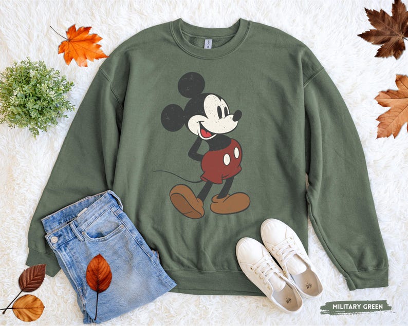 Puede incluir: Sudadera verde militar con un dise&ntilde;o de Mickey Mouse de estilo vintage. La parte superior de manga larga se combina con vaqueros azules y zapatillas blancas, creando un look casual. Hojas de oto&ntilde;o y una peque&ntilde;a planta a&ntilde;aden acentos de temporada.