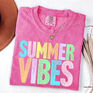 May include: A hot pink t-shirt with the words "SUMMER VIBES" in colorful, textured letters. A gold necklace with a pendant rests on the shirt. A brown hat and dried palm leaves are in the background.