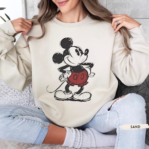 Puede incluir: Una sudadera de color arena con un gr&aacute;fico desgastado de Mickey Mouse en negro, rojo y blanco. El personaje de dibujos animados est&aacute; de pie con las manos en las caderas. La palabra "SAND" est&aacute; impresa en un rect&aacute;ngulo blanco.