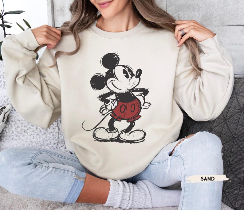 Puede incluir: Una sudadera de color arena con un gr&aacute;fico desgastado de Mickey Mouse en negro, rojo y blanco. El personaje de dibujos animados est&aacute; de pie con las manos en las caderas. La palabra "SAND" est&aacute; impresa en un rect&aacute;ngulo blanco.