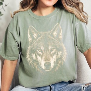 Puede incluir: Camiseta verde salvia con una ilustraci&oacute;n detallada de la cara de un lobo. El pelaje del lobo est&aacute; representado en tonos marrones y beige, con penetrantes ojos amarillos. La camiseta tiene un corte relajado y mangas cortas, adecuada para uso diario.