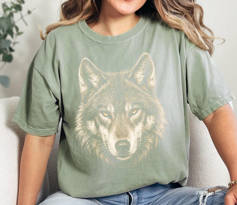 Puede incluir: Camiseta verde salvia con una ilustraci&oacute;n detallada de la cara de un lobo. El pelaje del lobo est&aacute; representado en tonos marrones y beige, con penetrantes ojos amarillos. La camiseta tiene un corte relajado y mangas cortas, adecuada para uso diario.
