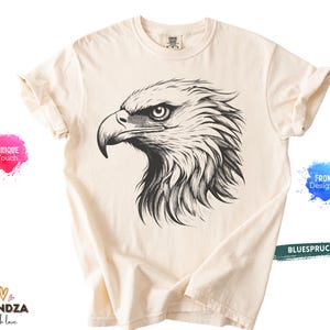 May include: Cream-colored t-shirt featuring a detailed black and white illustration of an eagle's head. The eagle's eye is focused, and the feathers are intricately drawn. The shirt has rolled-up sleeves and a crew neck.