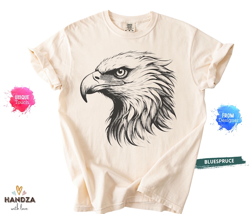 May include: Cream-colored t-shirt featuring a detailed black and white illustration of an eagle's head. The eagle's eye is focused, and the feathers are intricately drawn. The shirt has rolled-up sleeves and a crew neck.