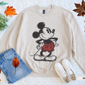 Puede incluir: Sudadera color arena con un gr&aacute;fico de Mickey Mouse en negro y rojo. La parte superior de manga larga se combina con vaqueros azules y zapatillas blancas. El dise&ntilde;o incluye el texto "SAND".