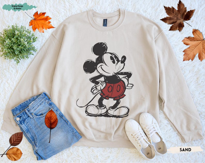Puede incluir: Sudadera color arena con un gr&aacute;fico de Mickey Mouse en negro y rojo. La parte superior de manga larga se combina con vaqueros azules y zapatillas blancas. El dise&ntilde;o incluye el texto "SAND".