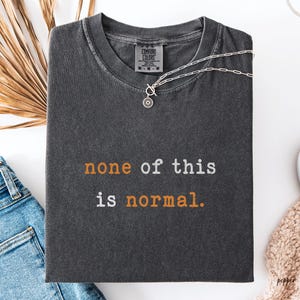 Peut inclure: T-shirt gris foncé Comfort Colors avec l'inscription "none of this is normal" en blanc et orange. Un collier argenté avec un pendentif rond est posé sur le t-shirt. Le t-shirt est plié sur une surface blanche.