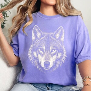 Puede incluir: Una camiseta de color lila con una ilustraci&oacute;n blanca detallada de la cara de un lobo. Los ojos del lobo est&aacute;n enfocados y el dise&ntilde;o est&aacute; centrado en la camiseta. La camiseta tiene mangas cortas y un corte relajado, adecuada para uso diario.