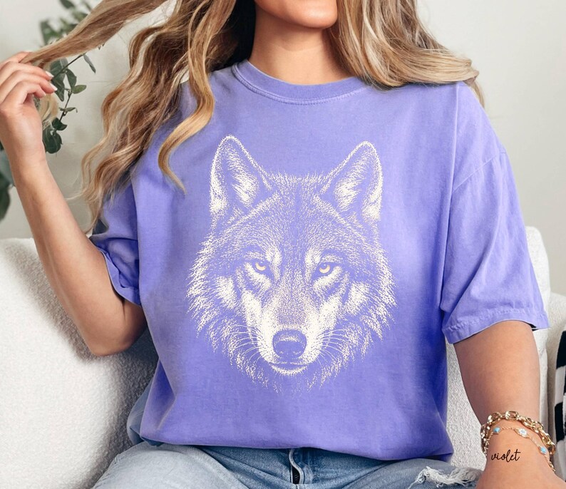 Puede incluir: Una camiseta de color lila con una ilustraci&oacute;n blanca detallada de la cara de un lobo. Los ojos del lobo est&aacute;n enfocados y el dise&ntilde;o est&aacute; centrado en la camiseta. La camiseta tiene mangas cortas y un corte relajado, adecuada para uso diario.