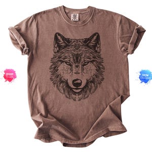 May include: Brown t-shirt featuring a detailed black illustration of a wolf's face. The shirt has a vintage, faded appearance and rolled-up sleeves. The wolf's fur is intricately drawn, with a focused gaze.