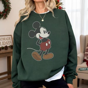 Puede incluir: Sudadera verde bosque con un dise&ntilde;o vintage de Mickey Mouse. El personaje de dibujos animados es de color negro, blanco, rojo y marr&oacute;n. La sudadera tiene cuello redondo y mangas largas. El texto "FOREST GREEN" es visible.