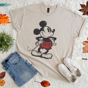 Puede incluir: Camiseta color arena con un dise&ntilde;o vintage de Mickey Mouse, con pantalones cortos rojos y guantes blancos. La camiseta se combina con vaqueros azules y zapatillas blancas. El texto "Original Design by Randia" es visible. La palabra "SAND" tambi&eacute;n es visible.