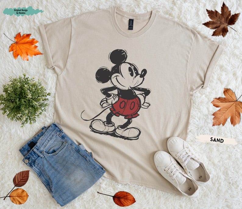 Puede incluir: Camiseta color arena con un dise&ntilde;o vintage de Mickey Mouse, con pantalones cortos rojos y guantes blancos. La camiseta se combina con vaqueros azules y zapatillas blancas. El texto "Original Design by Randia" es visible. La palabra "SAND" tambi&eacute;n es visible.