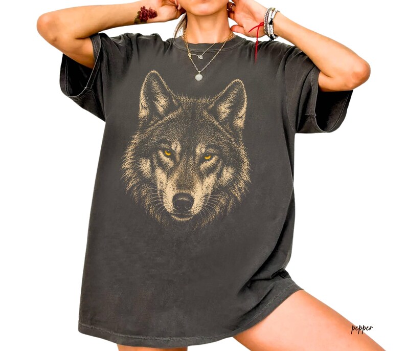 Puede incluir: Camiseta gris oscuro con una ilustraci&oacute;n detallada de la cara de un lobo con ojos amarillos. El dise&ntilde;o del lobo est&aacute; en tonos marrones y beige, sobre la tela gris oscuro. La camiseta tiene un corte relajado.