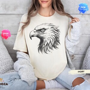 May include: Ivory-colored t-shirt featuring a detailed black and white illustration of an eagle's head. The design showcases the bird's sharp beak and intense gaze. The shirt is labeled "IVORY" and has the text "UNIQUE Touch" and "HANDZA with love".