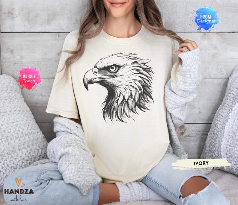 May include: Ivory-colored t-shirt featuring a detailed black and white illustration of an eagle's head. The design showcases the bird's sharp beak and intense gaze. The shirt is labeled "IVORY" and has the text "UNIQUE Touch" and "HANDZA with love".