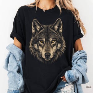 Puede incluir: Camiseta negra con una ilustraci&oacute;n detallada de la cara de un lobo. El lobo tiene ojos amarillos y un patr&oacute;n de pelaje detallado en tonos marrones y beige. La camiseta es de estilo cuello redondo cl&aacute;sico.