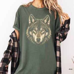 Puede incluir: Camiseta verde con una ilustraci&oacute;n detallada de la cara de un lobo. El lobo tiene ojos amarillos y un patr&oacute;n de pelaje detallado. La camiseta es de corte holgado. Se lleva una camisa a cuadros en blanco y negro encima.