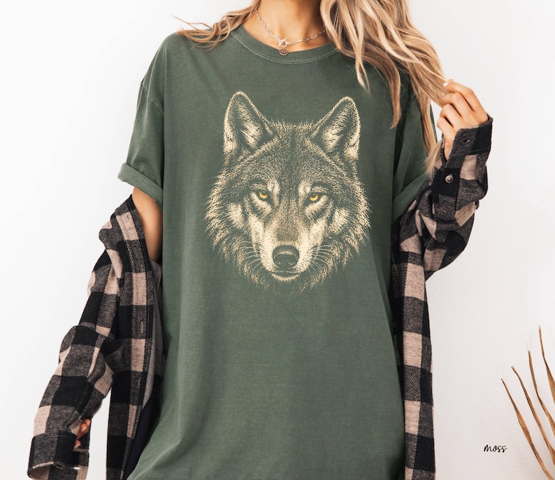 Puede incluir: Camiseta verde con una ilustraci&oacute;n detallada de la cara de un lobo. El lobo tiene ojos amarillos y un patr&oacute;n de pelaje detallado. La camiseta es de corte holgado. Se lleva una camisa a cuadros en blanco y negro encima.