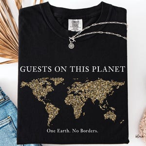 May include: Black t-shirt with the words "GUESTS ON THIS PLANET" and a gold pixelated world map design. The text "One Earth. No Borders." is below the map. A silver necklace with a circular pendant rests on the shirt.