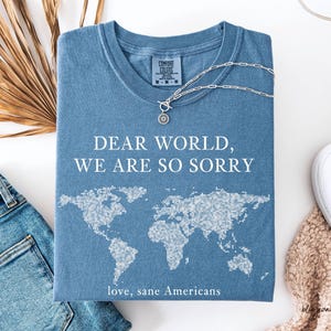 May include: A blue t-shirt with the text "DEAR WORLD, WE ARE SO SORRY" and a world map graphic. The shirt also includes the text "love, sane Americans". A silver necklace with a sun pendant is on the shirt.
