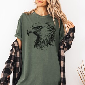 May include: Olive green t-shirt featuring a detailed black illustration of an eagle's head. The eagle's profile is prominently displayed, showcasing its sharp beak and intense gaze. The shirt is a casual style, suitable for everyday wear.
