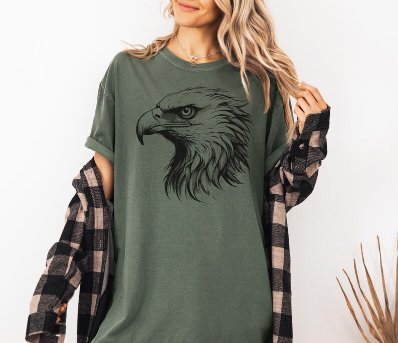 May include: Olive green t-shirt featuring a detailed black illustration of an eagle's head. The eagle's profile is prominently displayed, showcasing its sharp beak and intense gaze. The shirt is a casual style, suitable for everyday wear.