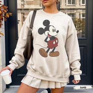 Puede incluir: Una sudadera de color arena con un gr&aacute;fico desgastado de Mickey Mouse. El personaje de dibujos animados est&aacute; representado en negro, rojo y marr&oacute;n. La sudadera tiene cuello redondo y mangas largas. La palabra "SAND" est&aacute; impresa en la parte inferior de la imagen.