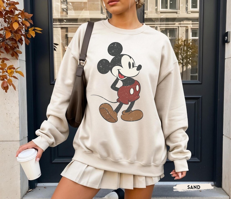 Puede incluir: Una sudadera de color arena con un gr&aacute;fico desgastado de Mickey Mouse. El personaje de dibujos animados est&aacute; representado en negro, rojo y marr&oacute;n. La sudadera tiene cuello redondo y mangas largas. La palabra "SAND" est&aacute; impresa en la parte inferior de la imagen.