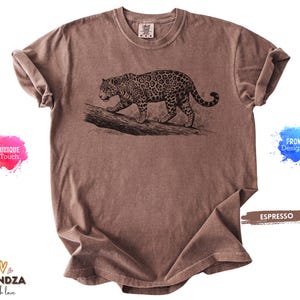 May include: A short-sleeved espresso brown t-shirt featuring a black ink-style illustration of a leopard walking along a fallen log. The shirt has a crew neckline and a relaxed fit, with the text "ESPRESSO" printed on a brown rectangular graphic.