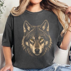 Puede incluir: Camiseta gris oscuro con una ilustraci&oacute;n detallada de la cara de un lobo. El lobo tiene ojos dorados y un dise&ntilde;o texturizado similar a la piel. La camiseta es de corte holgado, adecuada para uso casual.