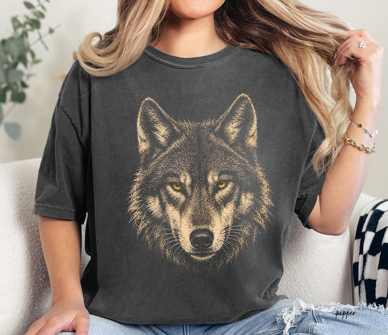 Puede incluir: Camiseta gris oscuro con una ilustraci&oacute;n detallada de la cara de un lobo. El lobo tiene ojos dorados y un dise&ntilde;o texturizado similar a la piel. La camiseta es de corte holgado, adecuada para uso casual.