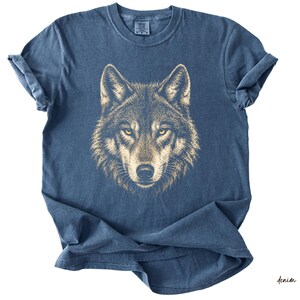 Puede incluir: Una camiseta azul denim con una ilustraci&oacute;n detallada de la cara de un lobo. El pelaje del lobo es de color marr&oacute;n claro y sus ojos son de un amarillo brillante. La camiseta tiene un cuello redondo cl&aacute;sico y mangas cortas.