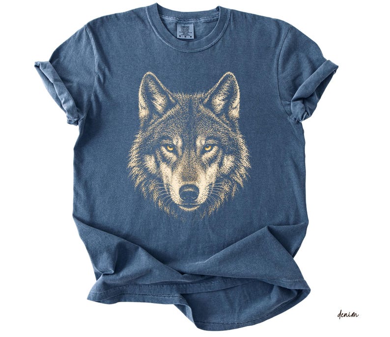 Puede incluir: Una camiseta azul denim con una ilustraci&oacute;n detallada de la cara de un lobo. El pelaje del lobo es de color marr&oacute;n claro y sus ojos son de un amarillo brillante. La camiseta tiene un cuello redondo cl&aacute;sico y mangas cortas.