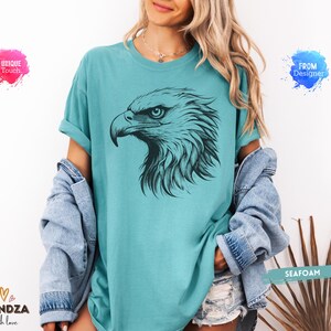 May include: Seafoam green t-shirt featuring a detailed black illustration of an eagle's head. The eagle's eye is visible, and the design is centered on the shirt. The shirt is part of a clothing collection.