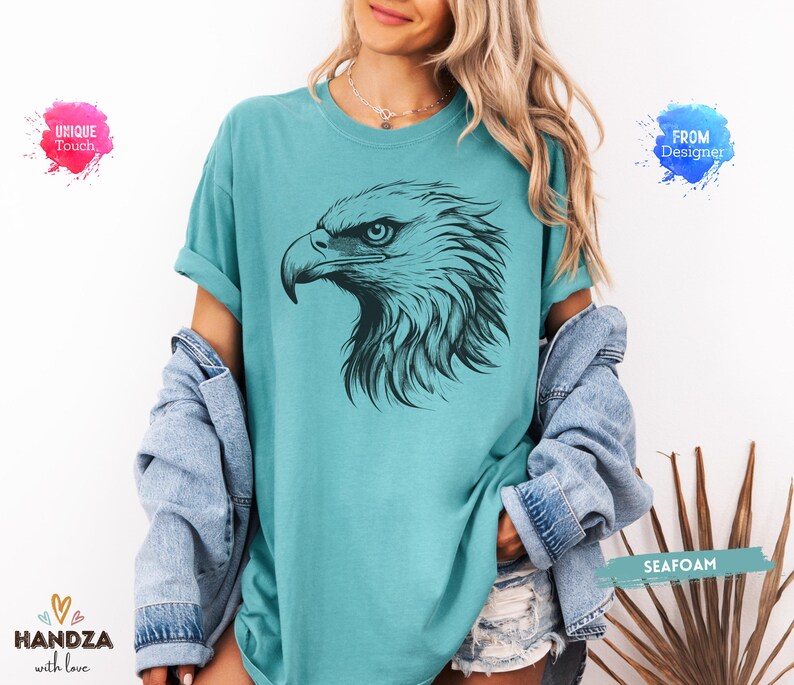 May include: Seafoam green t-shirt featuring a detailed black illustration of an eagle's head. The eagle's eye is visible, and the design is centered on the shirt. The shirt is part of a clothing collection.