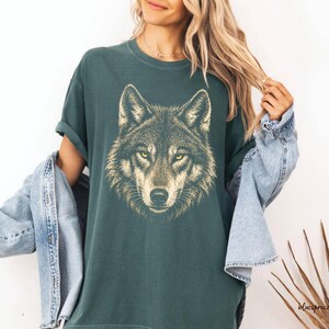 Puede incluir: Camiseta verde oscuro con una ilustraci&oacute;n detallada de la cabeza de un lobo. El pelaje del lobo est&aacute; representado en tonos marrones y beige, con ojos amarillos penetrantes. La camiseta tiene un corte relajado y mangas enrolladas.