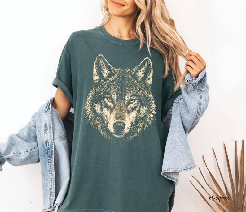 Puede incluir: Camiseta verde oscuro con una ilustraci&oacute;n detallada de la cabeza de un lobo. El pelaje del lobo est&aacute; representado en tonos marrones y beige, con ojos amarillos penetrantes. La camiseta tiene un corte relajado y mangas enrolladas.