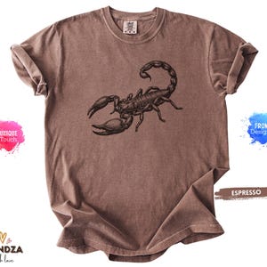 May include: Espresso-coloured t-shirt featuring a detailed black scorpion graphic. The shirt has a classic crew neck and short sleeves. The fabric appears soft and pre-shrunk. The shirt is a unique touch from the designer.