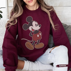 Puede incluir: Sudadera granate con un dise&ntilde;o de Mickey Mouse de estilo vintage. El personaje de dibujos animados se representa en su pose cl&aacute;sica, con pantalones cortos rojos y zapatos marrones. La palabra "MAROON" est&aacute; impresa en la esquina inferior derecha.