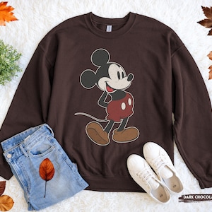 Puede incluir: Sudadera de color chocolate oscuro con un gr&aacute;fico de Mickey Mouse de estilo vintage. El personaje de dibujos animados est&aacute; representado en negro, rojo y marr&oacute;n. La sudadera se combina con vaqueros azules y zapatillas blancas, creando un look casual.