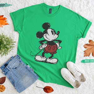 Puede incluir: Camiseta verde irland&eacute;s con un gr&aacute;fico desgastado de Mickey Mouse en negro, blanco y rojo. La camiseta se combina con vaqueros azules y zapatillas blancas. Hojas de oto&ntilde;o y una peque&ntilde;a planta est&aacute;n dispuestas alrededor de la camiseta.