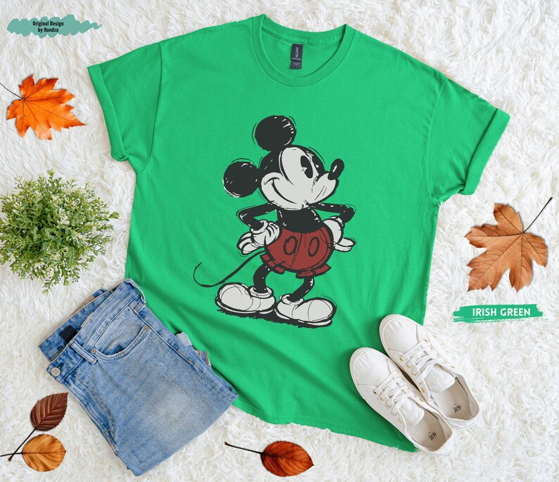 Puede incluir: Camiseta verde irland&eacute;s con un gr&aacute;fico desgastado de Mickey Mouse en negro, blanco y rojo. La camiseta se combina con vaqueros azules y zapatillas blancas. Hojas de oto&ntilde;o y una peque&ntilde;a planta est&aacute;n dispuestas alrededor de la camiseta.