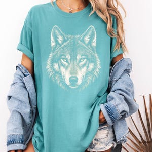 Puede incluir: Camiseta de color turquesa con un gr&aacute;fico detallado de la cara de un lobo blanco. La camiseta tiene mangas cortas y un corte holgado. El dise&ntilde;o del lobo est&aacute; centrado en la parte delantera, mostrando los ojos, la nariz y la textura del pelaje. Ropa casual.