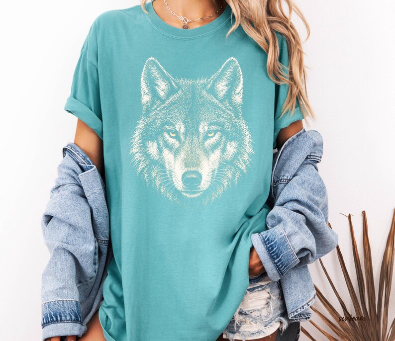 Puede incluir: Camiseta de color turquesa con un gr&aacute;fico detallado de la cara de un lobo blanco. La camiseta tiene mangas cortas y un corte holgado. El dise&ntilde;o del lobo est&aacute; centrado en la parte delantera, mostrando los ojos, la nariz y la textura del pelaje. Ropa casual.