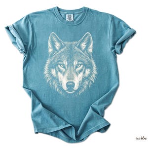 Puede incluir: Camiseta azul hielo con una ilustraci&oacute;n blanca de la cara de un lobo. El pelaje del lobo est&aacute; finamente detallado, con ojos enfocados y una nariz prominente. La camiseta es de color liso, con mangas enrolladas y cuello redondo.