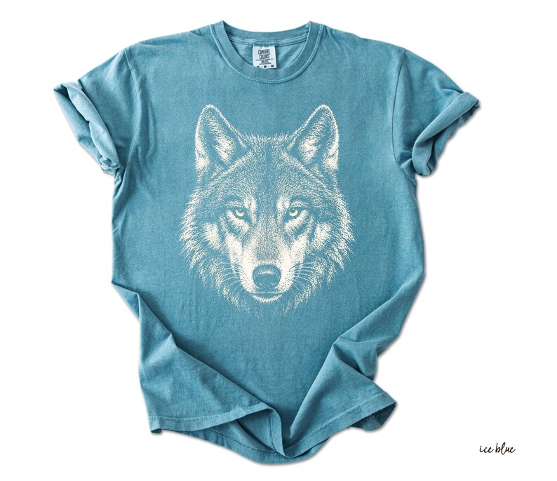 Puede incluir: Camiseta azul hielo con una ilustraci&oacute;n blanca de la cara de un lobo. El pelaje del lobo est&aacute; finamente detallado, con ojos enfocados y una nariz prominente. La camiseta es de color liso, con mangas enrolladas y cuello redondo.