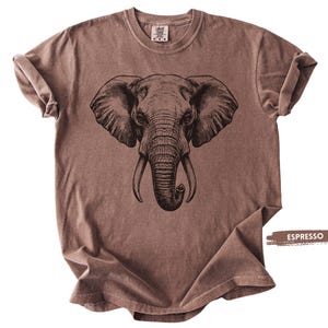 May include: Espresso-colored t-shirt featuring a detailed black illustration of an elephant's face. The elephant's large ears, tusks, and trunk are prominently displayed. The shirt has a classic crew neck and short sleeves.